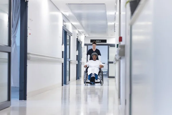 Diverse female doctor walking with senior female patient in wheelchair in hospital corridor. Medicine, healthcare and medical services, unaltered.