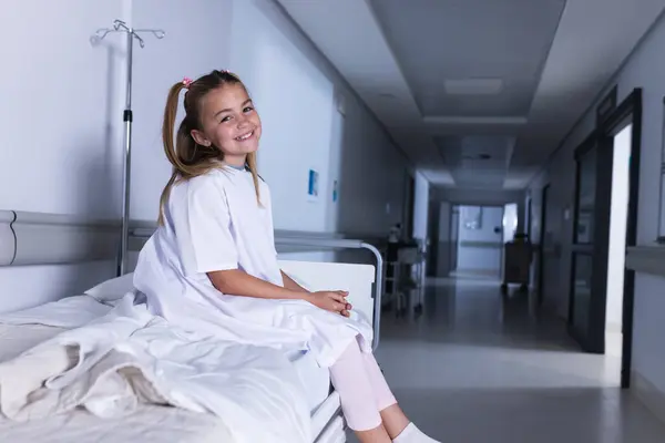 Happy caucasian girl sitting on bed in hospital corridor with copy space. Medicine, healthcare, childhood and medical services, unaltered.