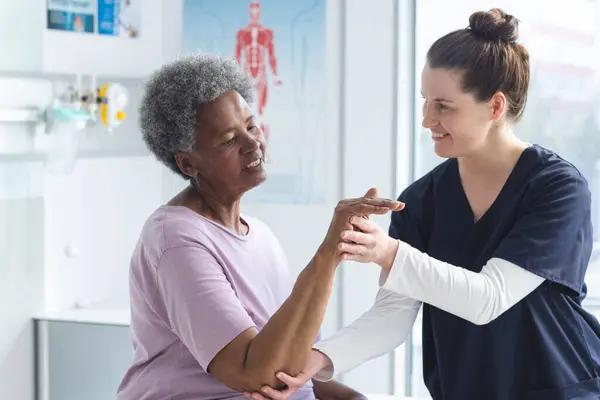 Diverse senior female patient exercising hand and female doctor advising in hospital room. Medicine, healthcare and medical services, unaltered.