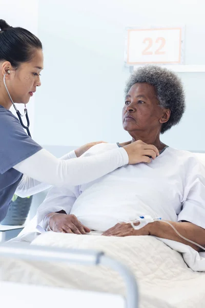 Diverse female doctor testing senior female patient using stethoscope in hospital room. Medicine, healthcare and medical services, unaltered.