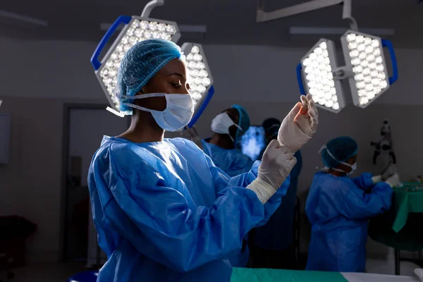 African american female doctor with face mask wearing protective gloves in hospital operating room. Medicine, healthcare and medical services, unaltered.