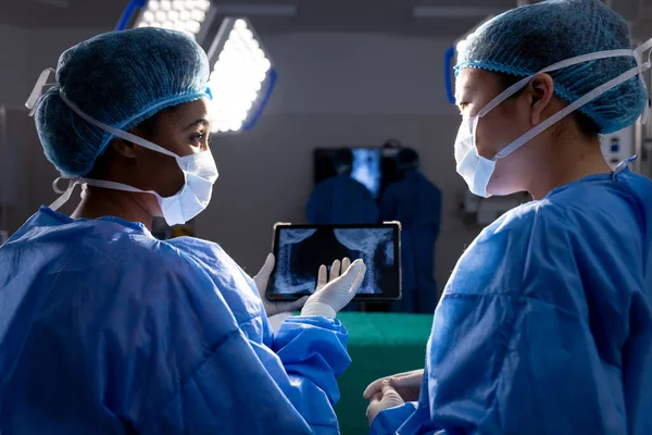 Diverse female doctors with face masks looking at x-ray on tablet in hospital operating room. Medicine, healthcare, communication and medical services, unaltered.