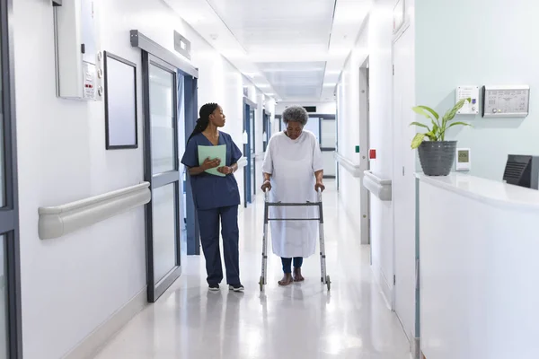 African american female doctor walking with senior female patient with crutches in hospital corridor. Medicine, healthcare and medical services, unaltered.
