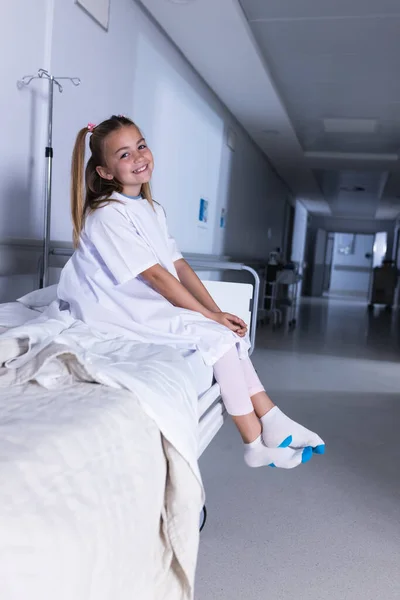 Happy caucasian girl sitting on bed in hospital corridor. Medicine, healthcare, childhood and medical services, unaltered.