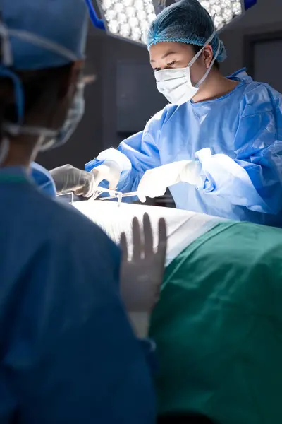 Diverse female doctor with face mask doing surgery in group of doctors in hospital operating room. Medicine, healthcare and medical services, unaltered.