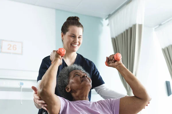 Diverse senior female patient exercising with weights and female doctor advising in hospital room. Medicine, healthcare and medical services, unaltered.