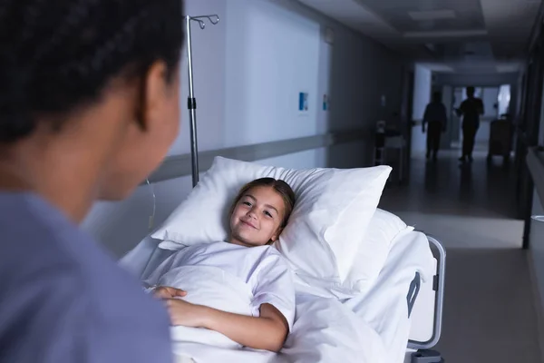 Happy diverse female doctor talking with girl lying in bed in hospital corridor. Medicine, healthcare, childhood and medical services, unaltered.