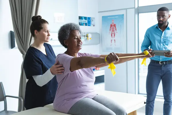 Diverse senior female patient exercising with band and female doctor advising in hospital room. Medicine, healthcare and medical services, unaltered.