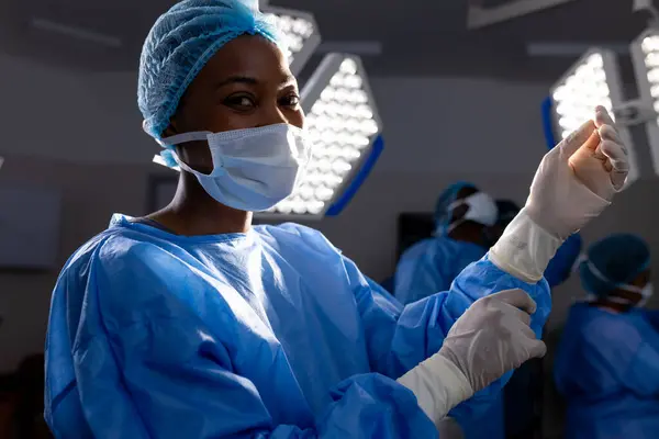 African american female doctor with face mask wearing protective gloves in hospital operating room. Medicine, healthcare and medical services, unaltered.