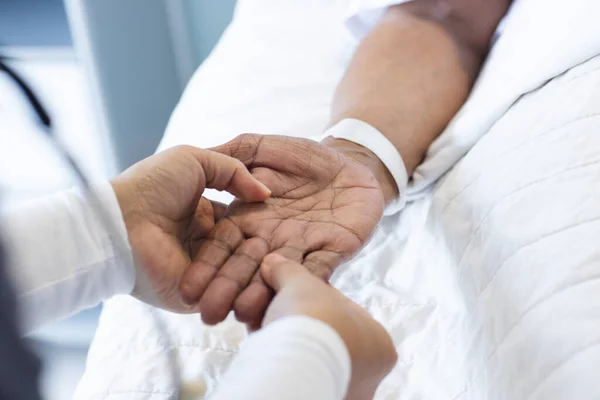 Hands of diverse female doctor holding hand of senior female patient in hospital room. Medicine, healthcare and medical services, unaltered.