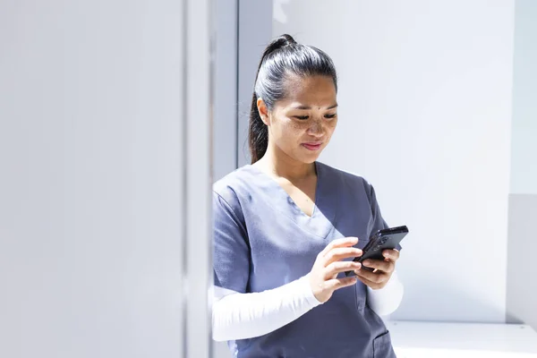 Biracial female doctor wearing scrubs using smartphon in hospital, copy space. Medicine, healthcare, communication, work and hospital, unaltered.