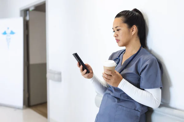 Médico Biracial Femenino Usando Matorrales Usando Smartphone Sosteniendo Café Para —  Fotos de Stock