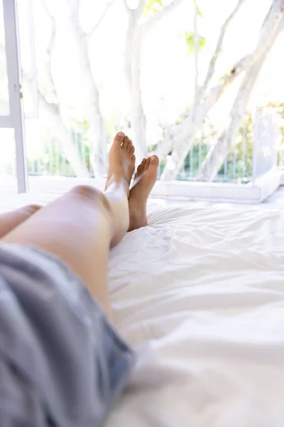 Legs of biracial woman lying on bed at sunny home. Lifestyle, free time and domestic life, unaltered.