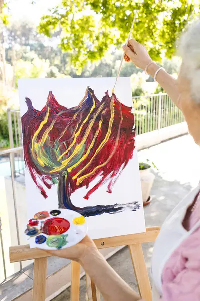 Caucasian senior woman painting on canvas on sunny terrace. Lifestyle, retirement, senior lifestyle, nature, creativity and domestic life, unaltered.