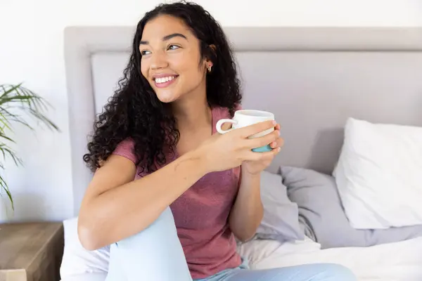 stock image Happy biracial woman holding cup of coffee sitting on bed at sunny home. Lifestyle, free time and domestic life, unaltered.