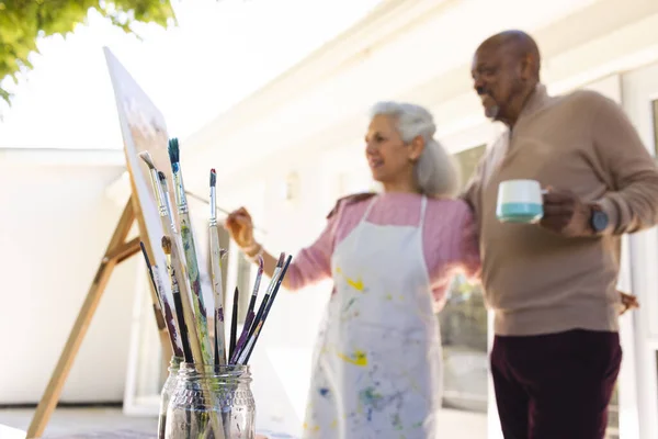 Diverse senior woman painting on canvas and senior man embracing her on sunny terrace. Lifestyle, retirement, senior lifestyle, nature, creativity, togetherness and domestic life, unaltered.