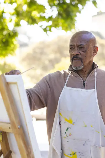 African american senior man painting on canvas on sunny terrace. Lifestyle, retirement, senior lifestyle, nature, creativity and domestic life, unaltered.