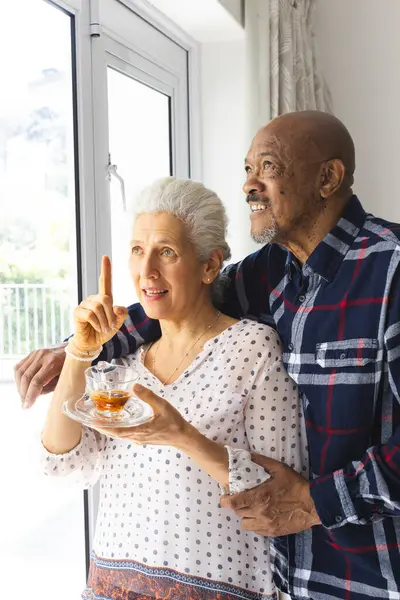Diverse senior couple drinking tea, embracing and looking out window at sunny home. Lifestyle, retirement, senior lifestyle, togetherness and domestic life, unaltered.