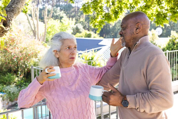 Diverse senior couple drinking coffee and talking on sunny terrace. Lifestyle, retirement, senior lifestyle, nature, togetherness and domestic life, unaltered.
