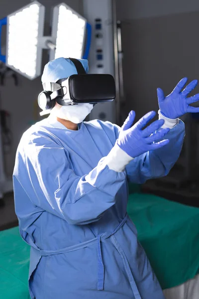 Biracial female surgeon wearing surgical gown using vr headset in operating theatre. Medicine, healthcare, surgery, technology, work and hospital, unaltered.