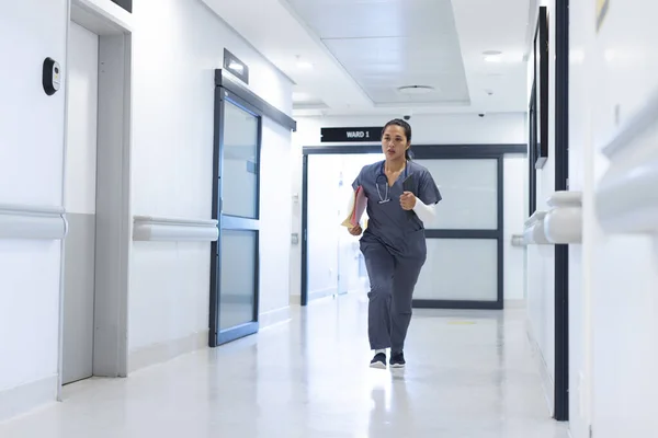 Biracial female doctor wearing scrubs running through corridor in hospital, copy space. Medicine, healthcare, work and hospital, unaltered.