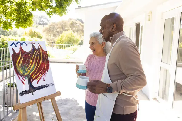 Happy diverse senior couple drinking coffee, embracing and looking at painting on sunny terrace. Lifestyle, retirement, senior lifestyle, nature, creativity, togetherness and domestic life, unaltered.