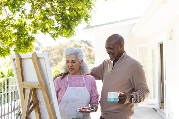 Happy diverse senior couple drinking coffee, embracing and looking at painting on sunny terrace. Lifestyle, retirement, senior lifestyle, nature, creativity, togetherness and domestic life, unaltered.