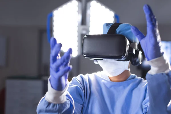 Biracial female surgeon wearing surgical gown using vr headset in operating theatre. Medicine, healthcare, surgery, technology, work and hospital, unaltered.
