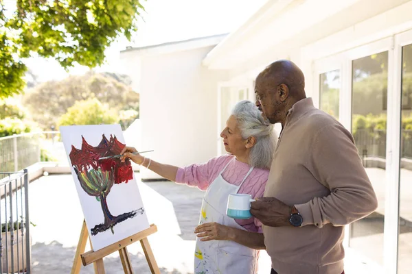 Diverse senior woman painting on canvas and senior man holding cup of coffee on sunny terrace. Lifestyle, retirement, senior lifestyle, nature, creativity, togetherness and domestic life, unaltered.