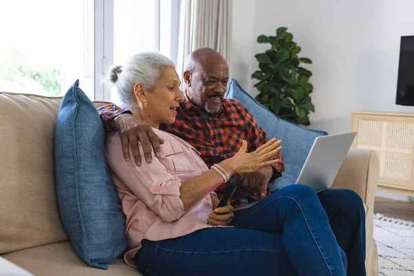 Diverse senior couple sitting on sofa, embracing and using laptop in sunny living room. Lifestyle, retirement, senior lifestyle, togetherness, communication and domestic life, unaltered.