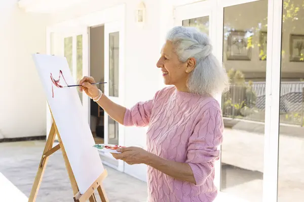 Happy caucasian senior woman painting on canvas on sunny terrace. Lifestyle, retirement, senior lifestyle, creativity and domestic life, unaltered.