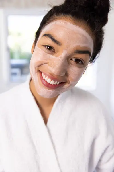 Portrait of happy biracial woman in bathrobe with face mask in sunny bathroom. Lifestyle, self care, beauty, skin and domestic life, unaltered.