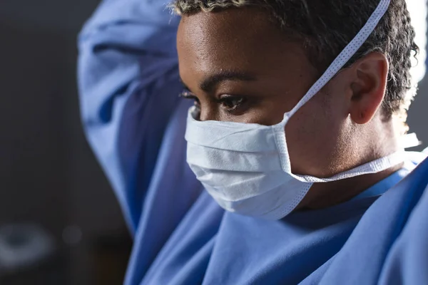 Biracial female surgeon wearing surgical gown and face mask in operating theatre. Medicine, healthcare, surgery, work and hospital, unaltered.