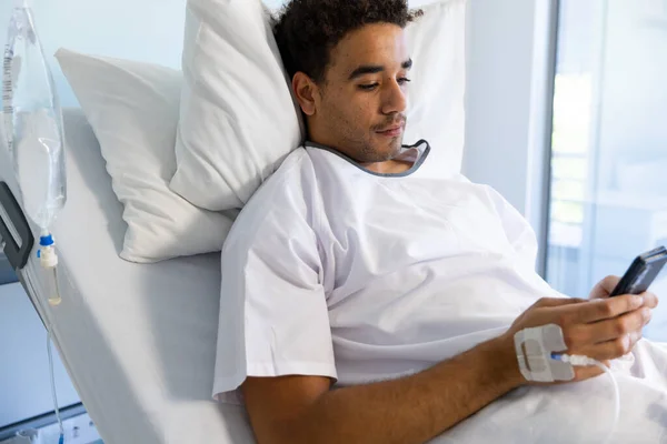 Biracial male patient using smartphone in bed with iv drip in sunny hospital room. Medicine, healthcare, communication and medical services, unaltered.