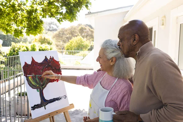 Happy diverse senior woman painting on canvas and senior man embracing her on sunny terrace. Lifestyle, retirement, senior lifestyle, nature, creativity, togetherness and domestic life, unaltered.