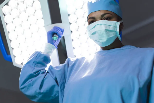 Portrait of biracial female surgeon wearing surgical gown and face mask in operating theatre. Medicine, healthcare, surgery, work and hospital, unaltered.