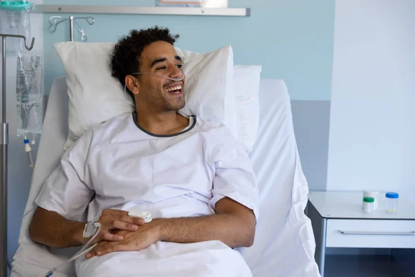 Happy biracial male patient in bed with iv drip in sunny hospital room. Medicine, healthcare and medical services, unaltered.