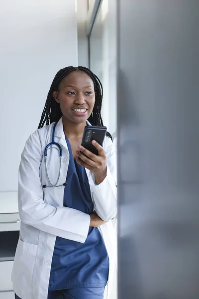 African american female doctor wearing lab coat using smartphone in hospital, copy space. Medicine, healthcare, communication, work and hospital, unaltered.