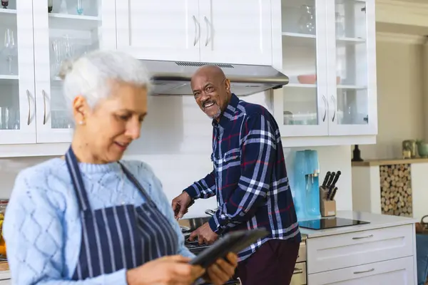 Diverse senior couple preparing meal using tablet in kitchen. Lifestyle, retirement, senior lifestyle, food, cooking, togetherness, communication, recipe and domestic life, unaltered.