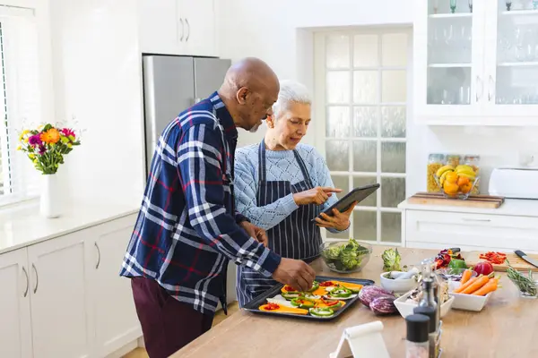 Diverse senior couple preparing meal using tablet in kitchen. Lifestyle, retirement, senior lifestyle, food, cooking, togetherness, communication, recipe and domestic life, unaltered.