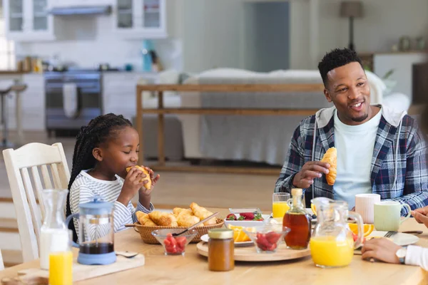 Happy african american girl eating croissant at table at home, copy space. Togetherness, parenthood, childhood, food and drink, domestic life and healthy lifestyle, unaltered.