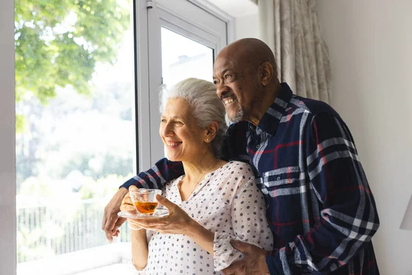 Happy diverse senior couple drinking tea, embracing and looking out window at sunny home. Lifestyle, retirement, senior lifestyle, togetherness and domestic life, unaltered.