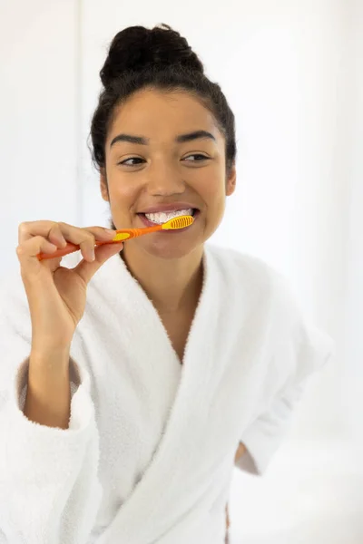 Happy biracial woman in bathrobe brushing teeth in sunny bathroom. Lifestyle, self care, hygiene and domestic life, unaltered.