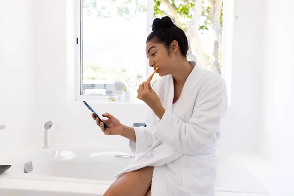 Biracial woman in bathrobe brushing teeth and using smartphone in sunny bathroom. Lifestyle, self care, hygiene, communication and domestic life, unaltered.