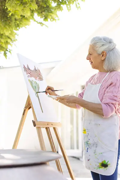 Caucasian senior woman painting on canvas on sunny terrace. Lifestyle, retirement, senior lifestyle, nature, creativity and domestic life, unaltered.