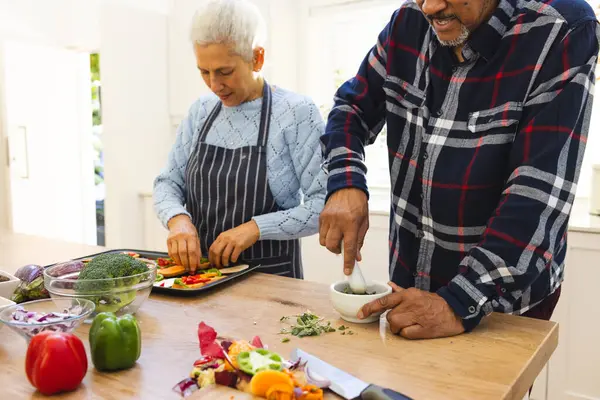 Diverse senior couple preparing healthy meal with vegetables in kitchen. Lifestyle, retirement, senior lifestyle, food, cooking, togetherness and domestic life, unaltered.