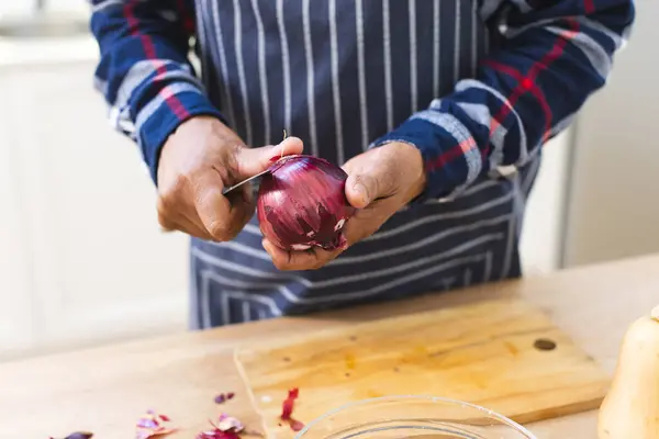 Hands of african american senior man in apron peeling onions in kitchen. Lifestyle, retirement, senior lifestyle, food, cooking and domestic life, unaltered.