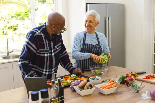 Diverse senior couple preparing healthy meal with vegetables using tablet in kitchen. Lifestyle, retirement, senior lifestyle, food, cooking, togetherness, recipe and domestic life, unaltered.