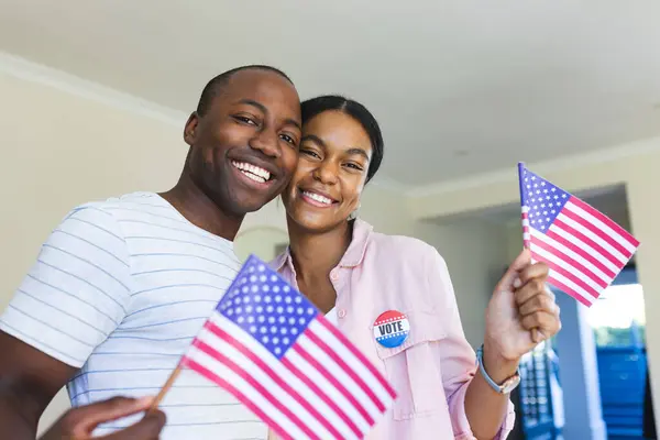 Young African American man and biracial woman holding flags at home, proudly displaying their \'I Voted\' stickers and vote badges. They show civic pride, celebrating democracy.