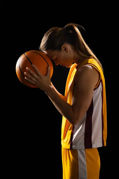 Focused young Caucasian female basketball player holds a basketball on a black background. She\'s in a sports uniform, portraying determination and concentration before a game.